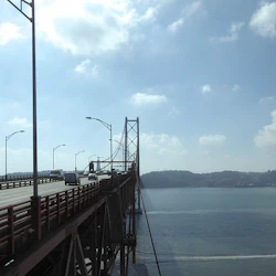 Bilar som kör på en hängbro över en lugn vattenmassa under en delvis molnig himmel. En båt lämnar en vaken i vattnet nedanför.