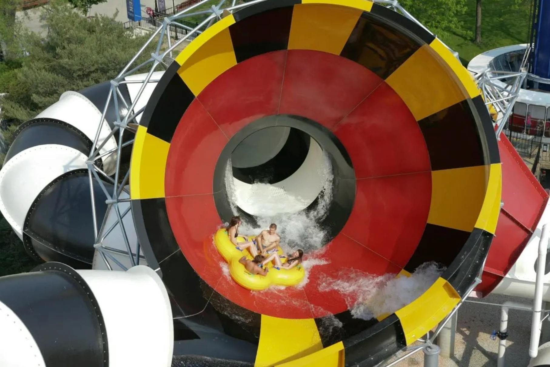 Quatre persones en una bassa inflable groga que surten d'un tobogan d'aigua de colors amb patrons vermells, grocs i negres.