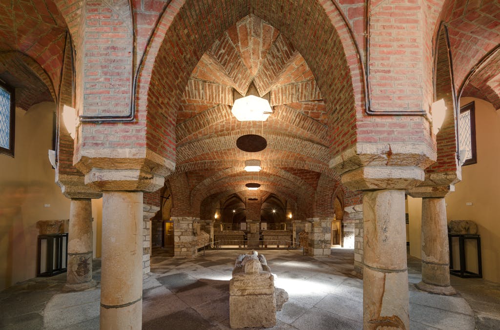 L'image montre une crypte voûtée en brique et en pierre avec des colonnes, éclairée par des lampes suspendues.