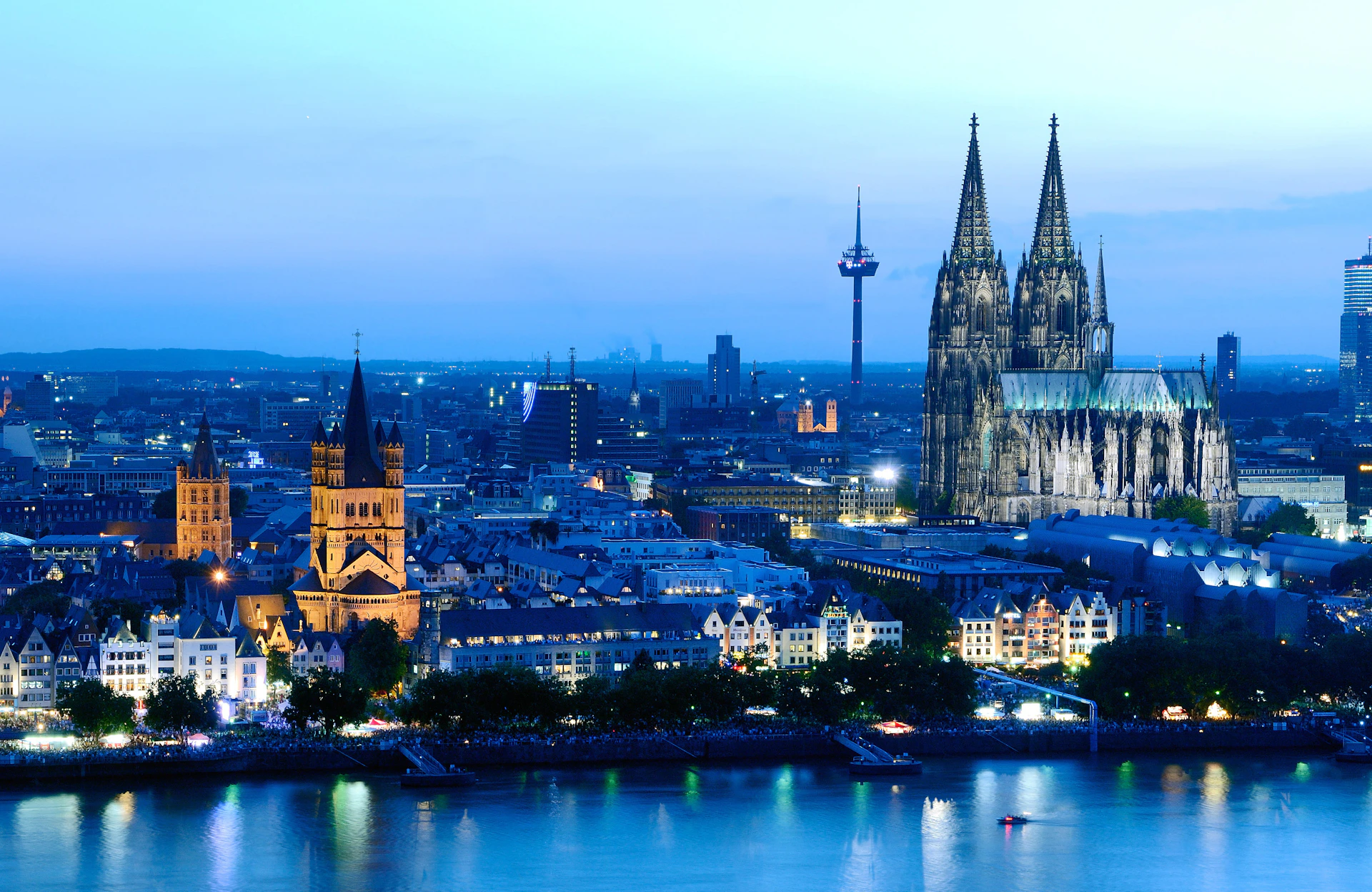 Aerial view of Cologne at dusk, featuring the illuminated Cologne Cathedral, St. Martin's Church, and the Rhine River in the foreground.