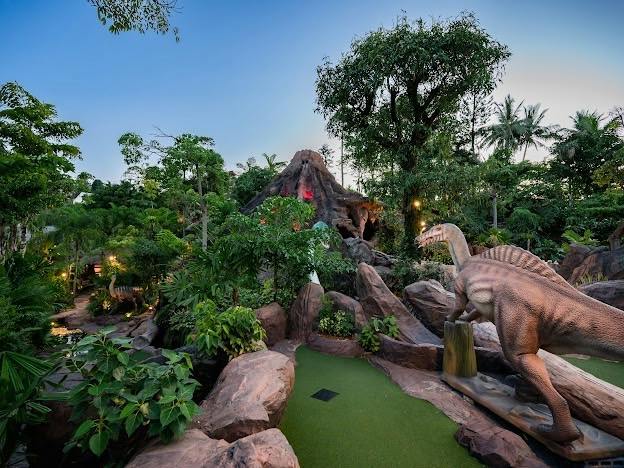 A miniature golf course with a dinosaur theme, featuring a dinosaur statue, lush greenery, and a volcano centerpiece.