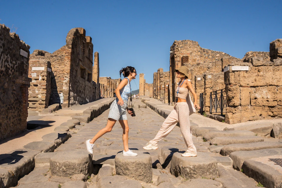 Two women in casual summer outfits stand and leap on large stone blocks in an ancient, ruined city under a clear blue sky.
