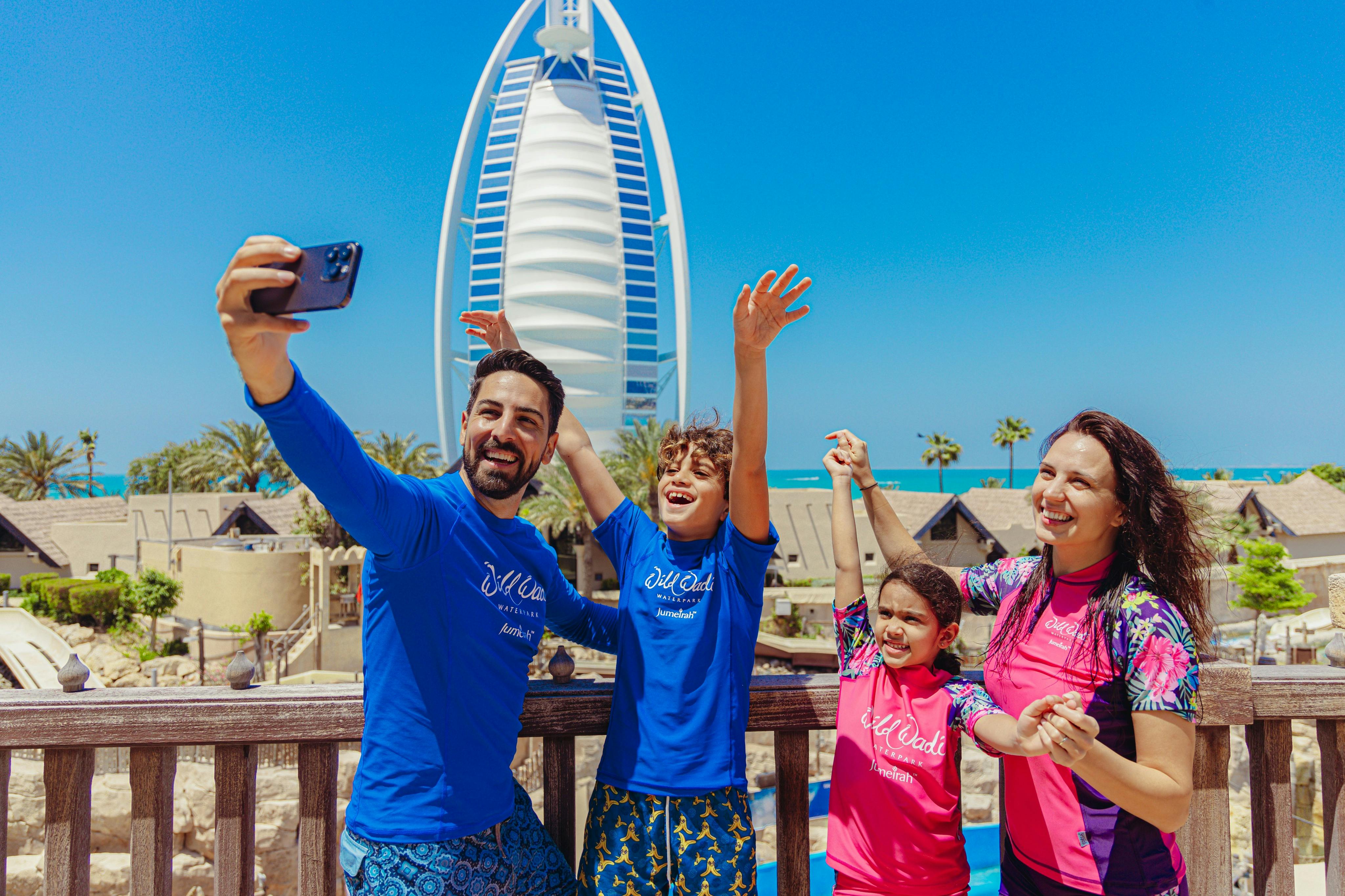 Une famille de quatre personnes souriant et posant pour une photo, avec en arrière-plan un bâtiment en forme de voile et un ciel d'un bleu éclatant.