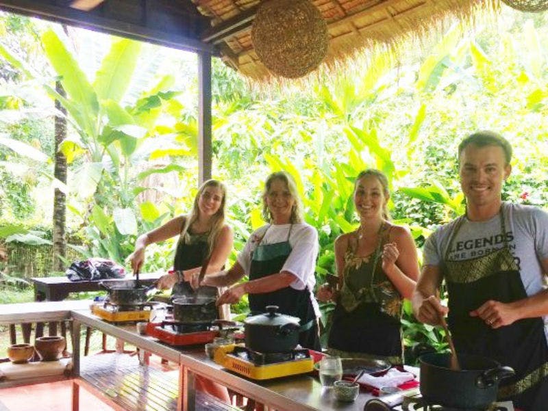 Cuatro personas sonriendo y cocinando en una barra de cocina rodeada de exuberante vegetación tropical.