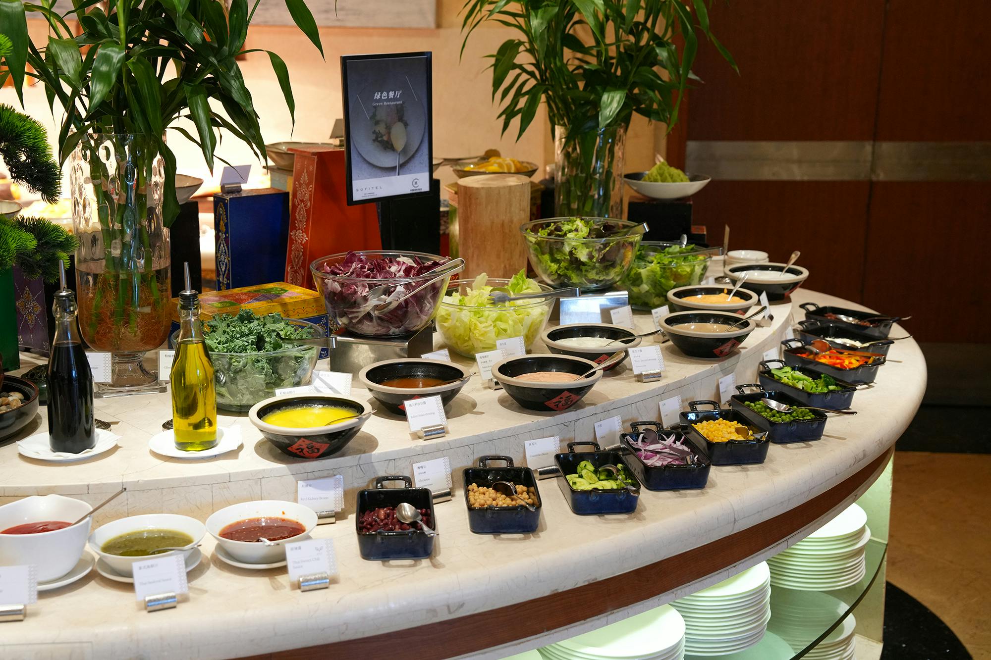 A salad bar setup with various vegetables, toppings, and dressings in bowls and containers, with leafy plants in the background.