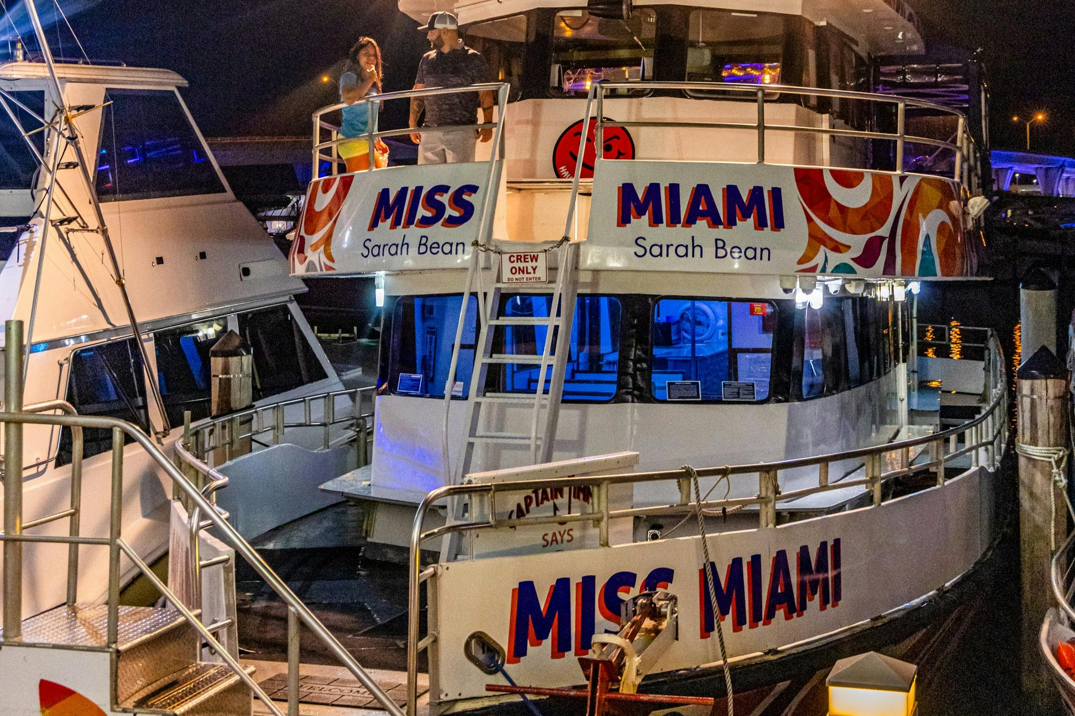 A boat named 'Miss Miami' docked at night, with passengers on the upper deck and illuminated interior spaces.