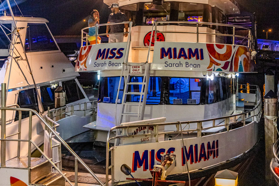 A boat named 'Miss Miami' docked at night, with passengers on the upper deck and illuminated interior spaces.
