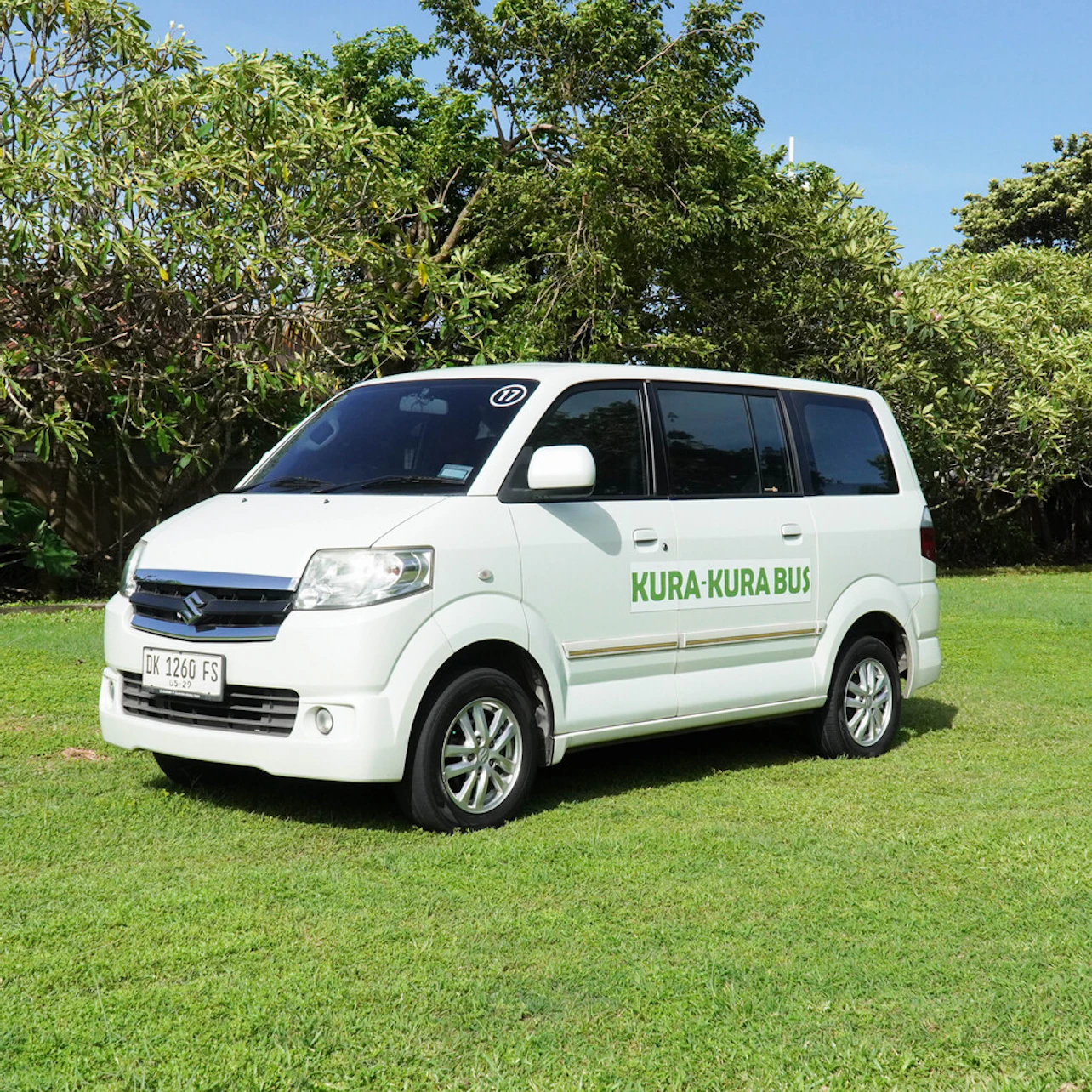A white van with "Kura-Kura Bus" written on the side, parked on a grassy field with trees in the background.