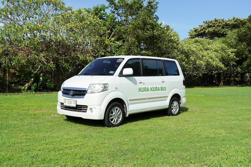 A white van with "Kura-Kura Bus" written on the side, parked on a grassy field with trees in the background.