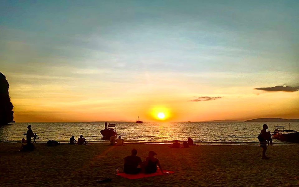 People sitting on a beach at sunset watching boats on the water. The sky is orange and blue.
