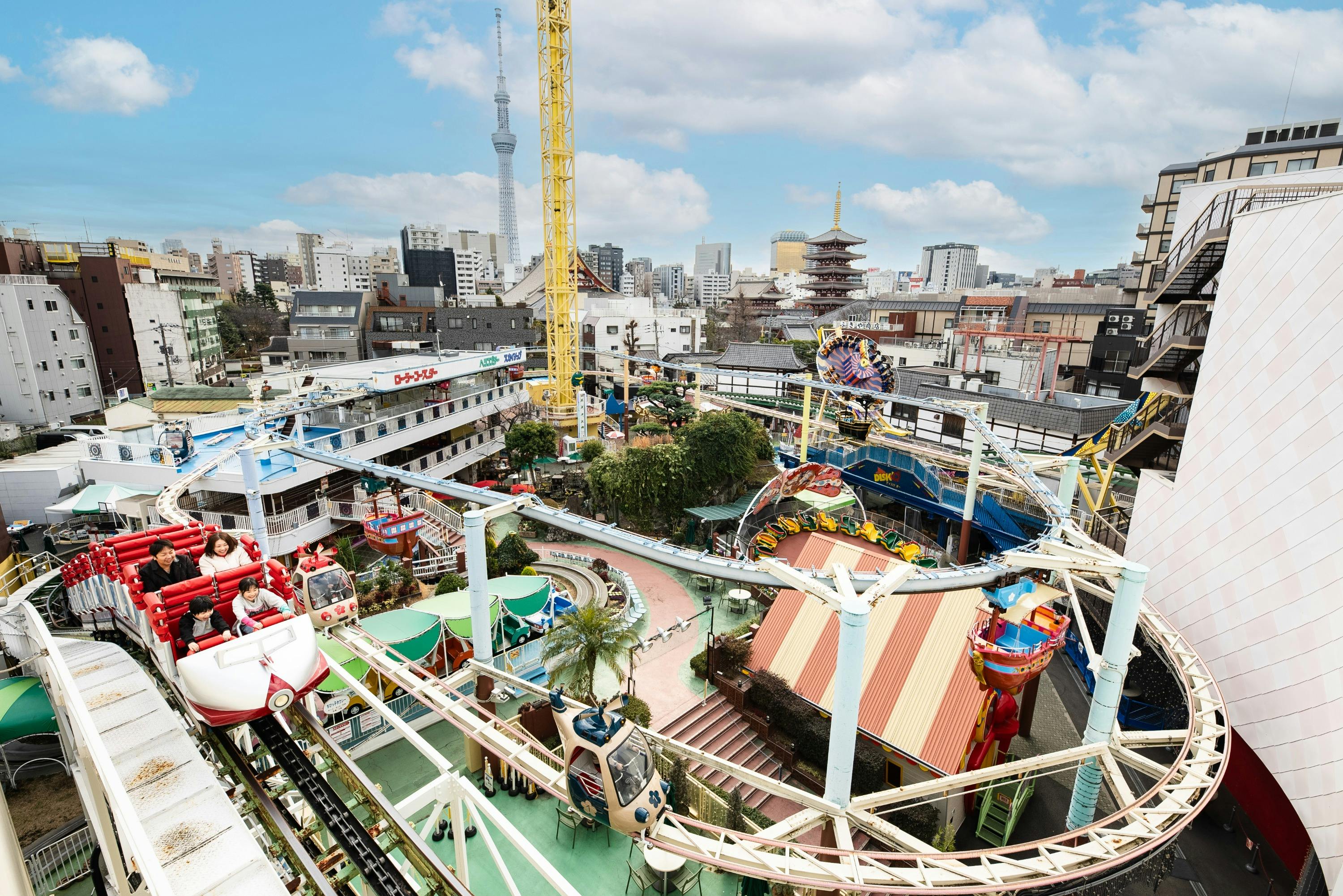 A colorful amusement park with rides and attractions, surrounded by an urban skyline and a blue sky with clouds.