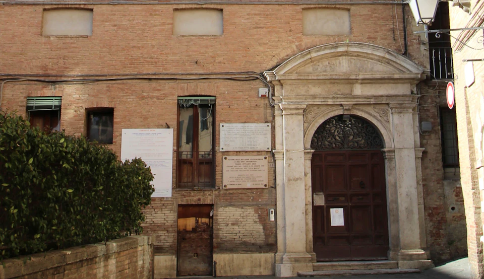 Brick building exterior with a large, arched wooden door, multiple plaques, and a small window above a wooden panel door.