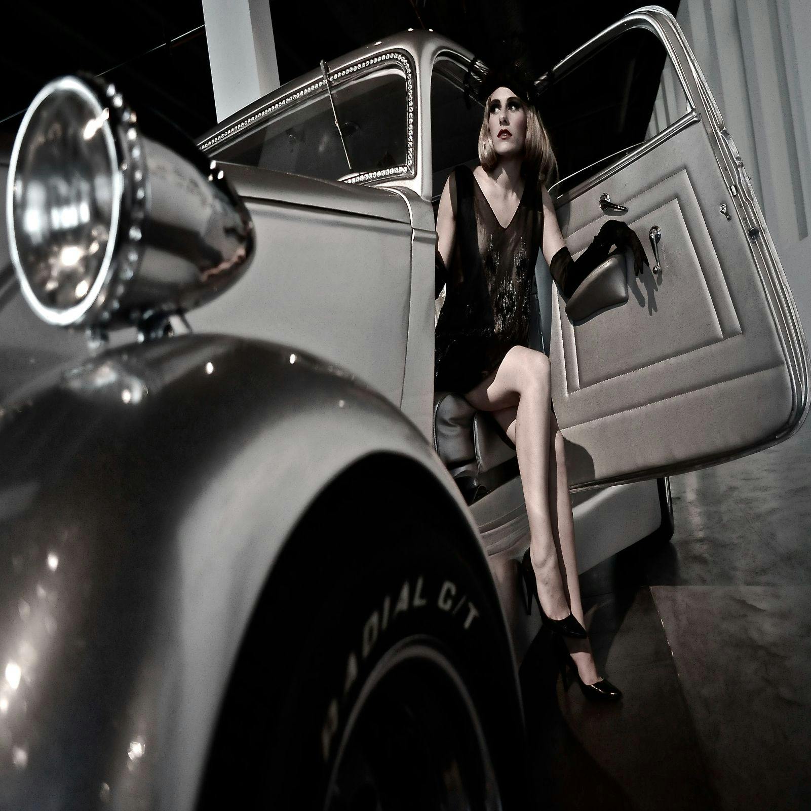 En kvinde i en sort kjole og handsker sidder i en åben veteranbil, der ser ud til at træde ud, med en forlygte og dæk i forgrunden.