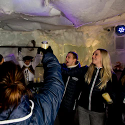 Människor i vinterjackor lyfter koppar för att fira inuti en isbar, med en bartender utklädd till pirat i bakgrunden.