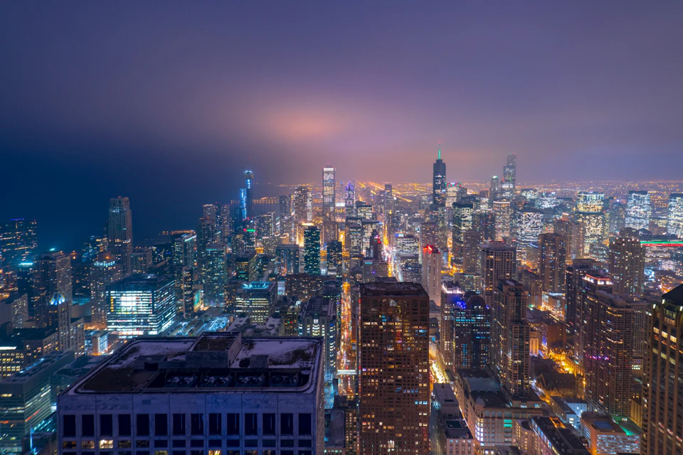 Aerial view of a city skyline at night with illuminated skyscrapers and buildings under a hazy purple sky.