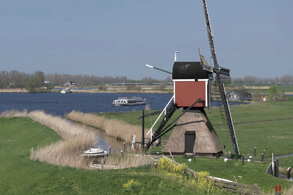A traditional windmill by a canal with reeds, a boat, and a river in the background, set in a green rural landscape.