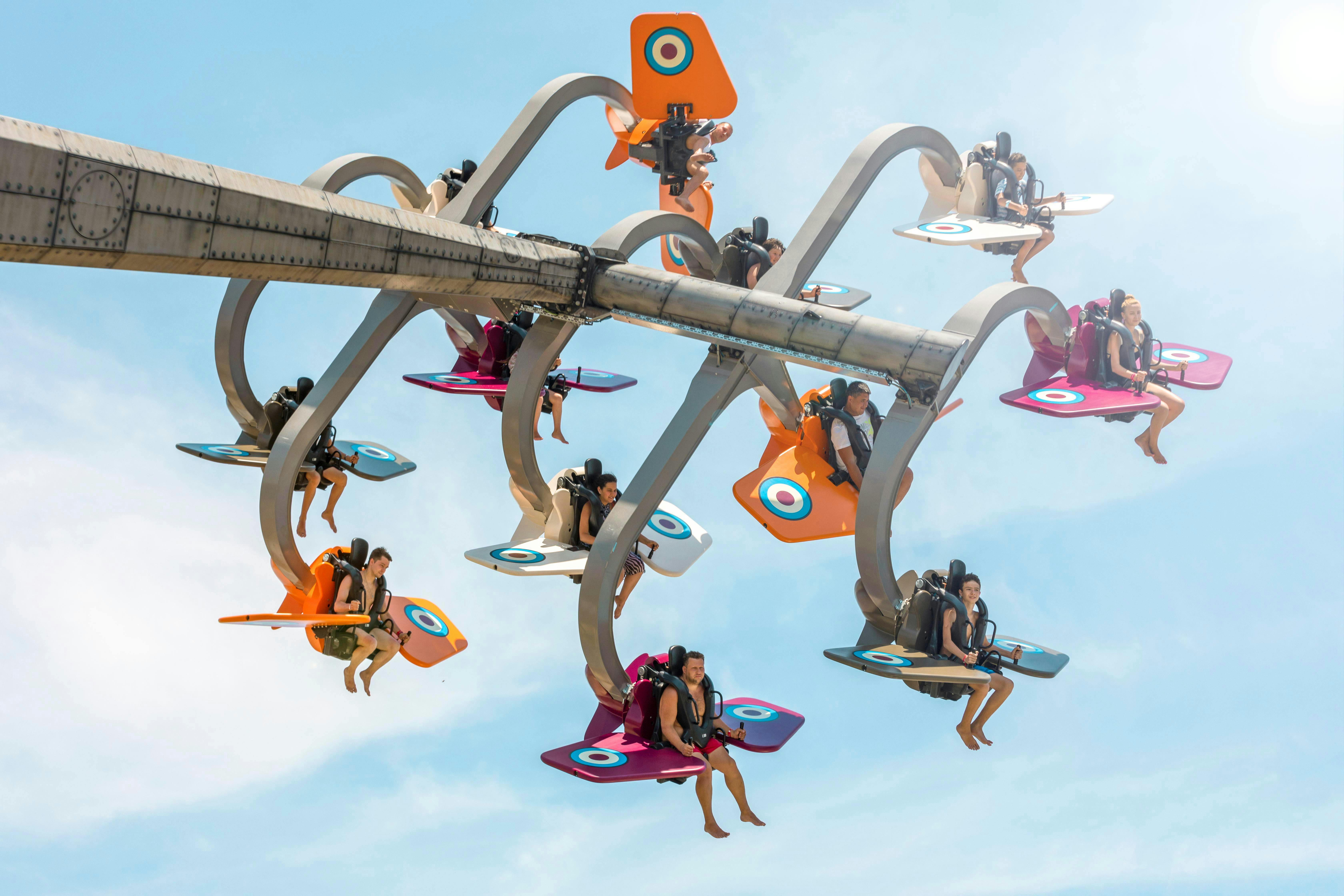 Amusement park ride with people seated on colorful wing-shaped carriers, suspended in the air, under a sunny sky.
