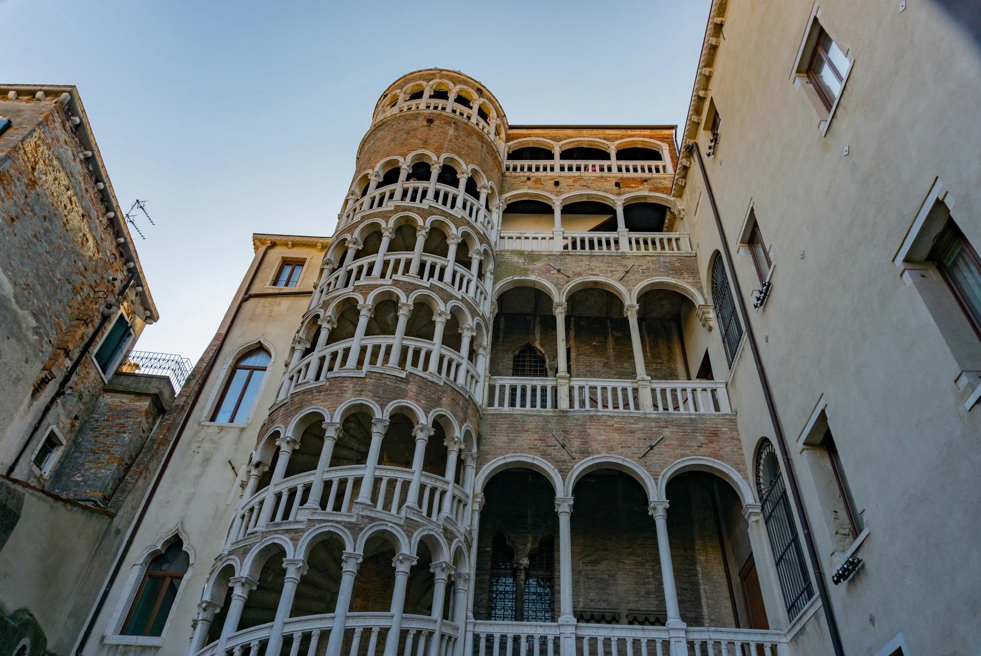 A multilevel spiral staircase with arched windows and a brick façade in a historic building against a clear sky.