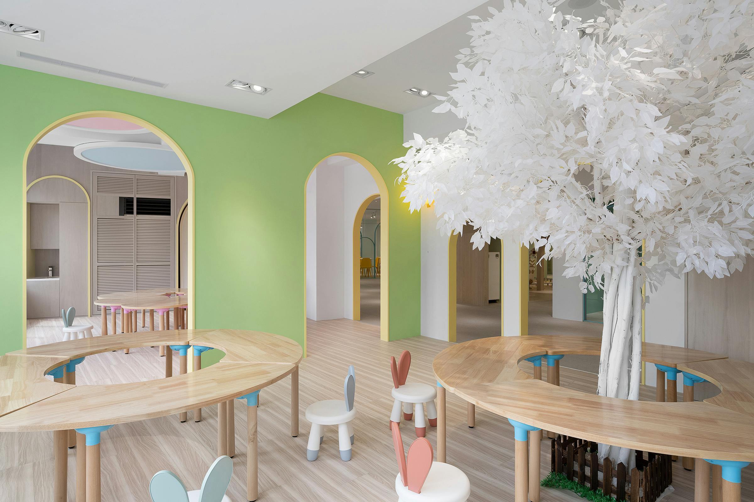A bright, modern children's playroom with wooden tables, colorful chairs, and a decorative white tree.