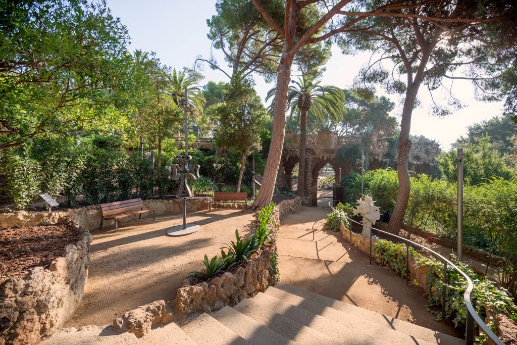 石段、ベンチ、彫刻、高い木やヤシの木などの緑豊かな緑が広がる日当たりの良い公園エリア。