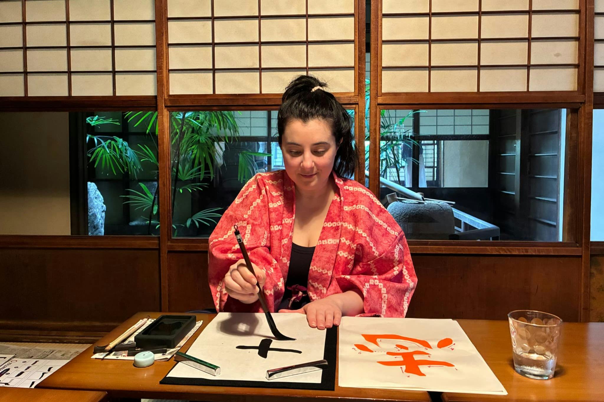 A woman wearing a pink kimono practices calligraphy at a table, with brushes, ink, and paper in a traditional room.