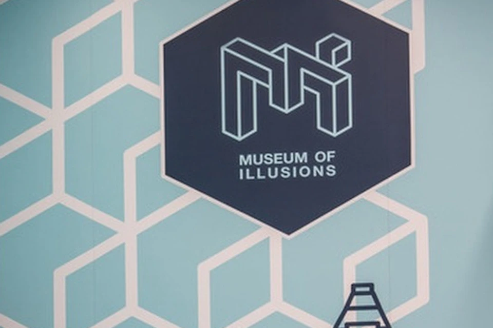 Two people in a room with geometric wall patterns and a "Museum of Illusions" logo, creating a perspective illusion.