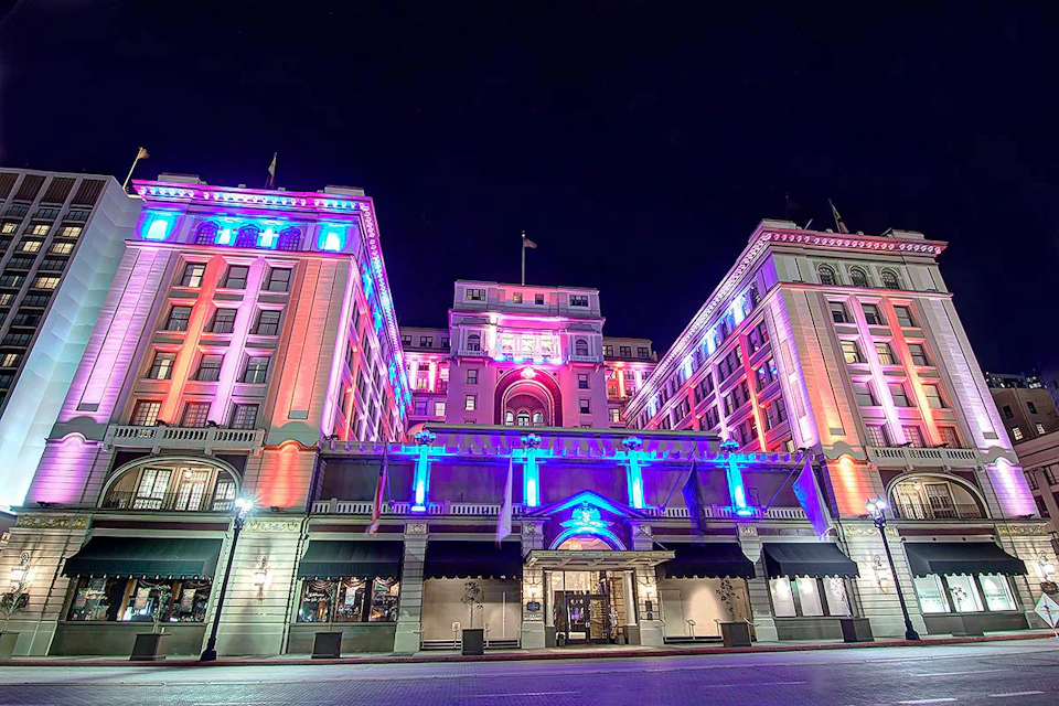 A large, ornate building lit with colorful lights (purple, red, and blue) against a dark night sky, featuring arched windows and awnings.