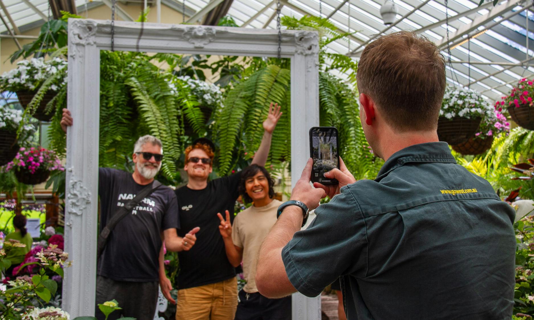 Trzy osoby pozują w dużej ramce w otoczeniu bujnej zieleni, a osoba robi im zdjęcie smartfonem.