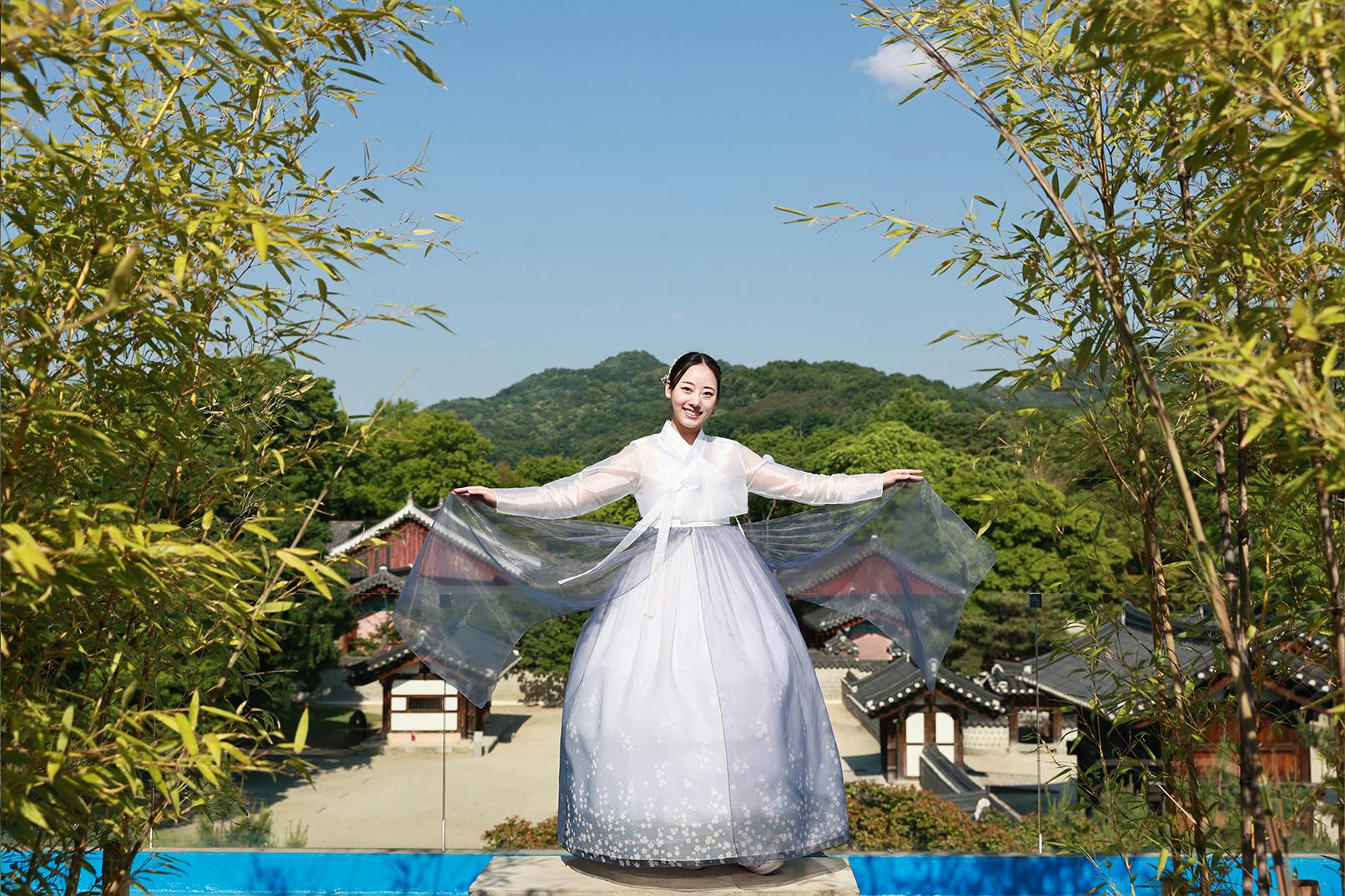 A person wearing a traditional white and lavender Korean hanbok stands in a garden with a backdrop of trees and traditional buildings.