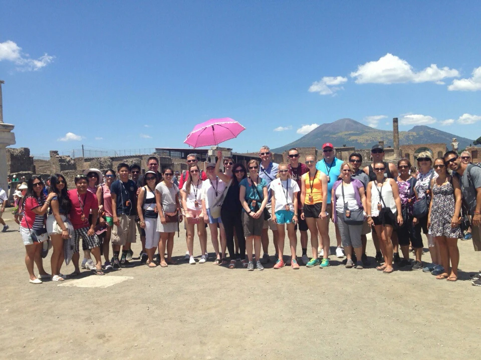 Group of people standing outdoors under a clear sky, with some holding umbrellas. Mountains and ancient ruins in the background.