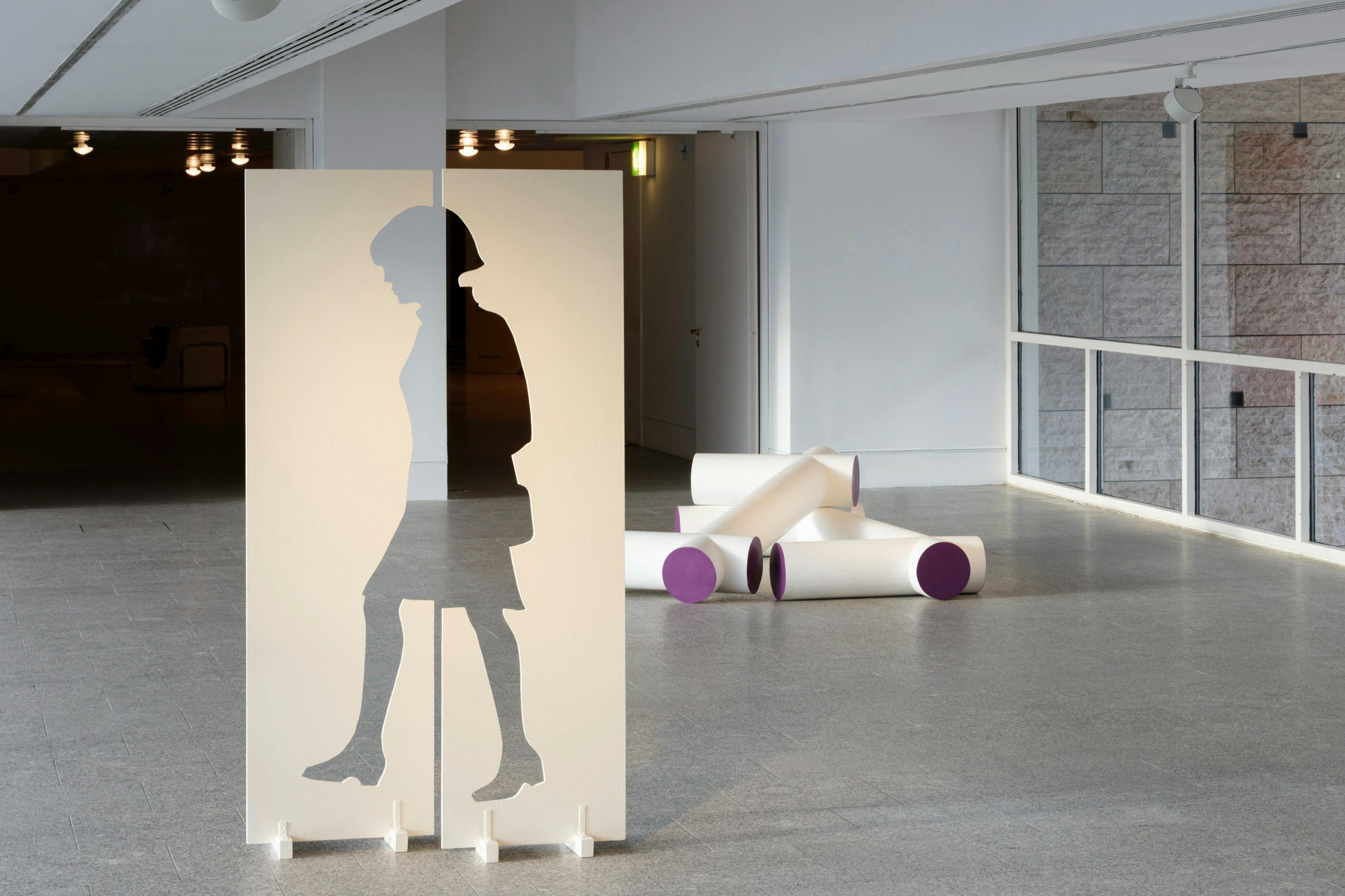Konstinstallation med en utskuren silhuettpanel och vita cylindriska former med lila ändar i ett modernt galleriutrymme.
