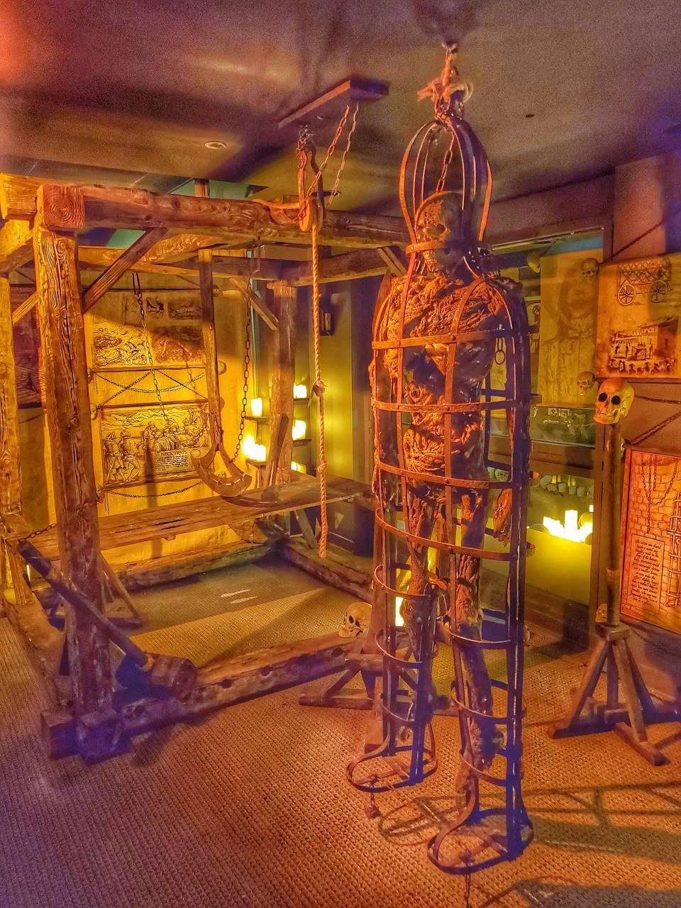 A medieval torture chamber display with a wooden rack, a metal cage containing a skeleton, and maps on the walls.