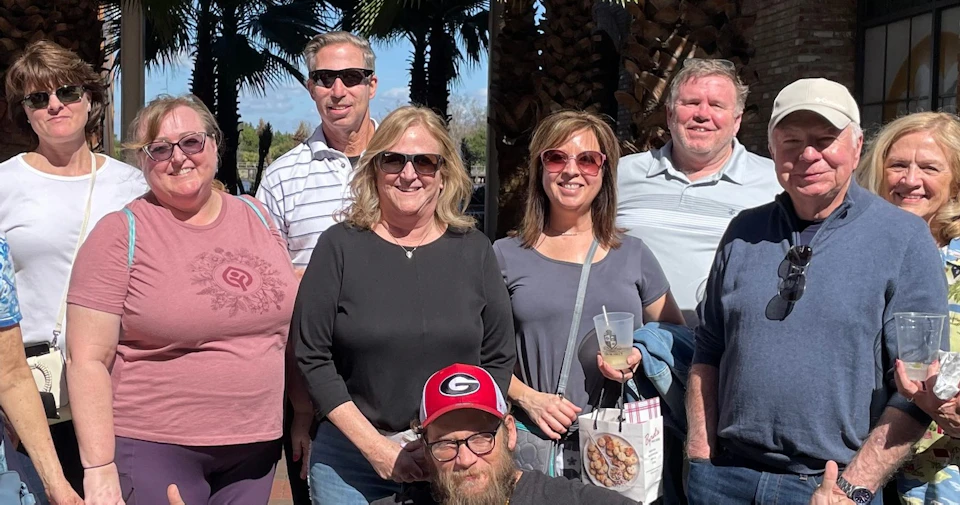 A group of people posing outdoors in a sunny setting with palm trees in the background. One person kneels in the foreground.