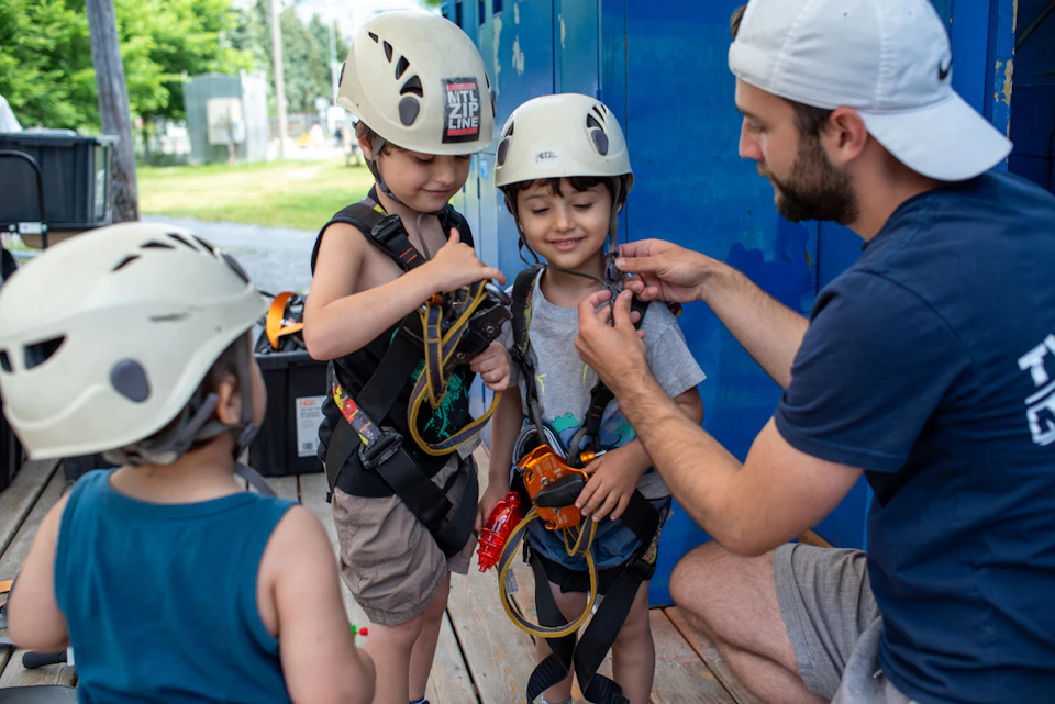 An adult helps two children in helmets and harnesses prepare for a zip lining activity near a blue storage area.