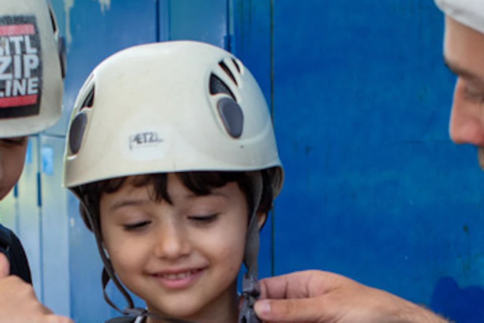 An adult helps two children in helmets and harnesses prepare for a zip lining activity near a blue storage area.