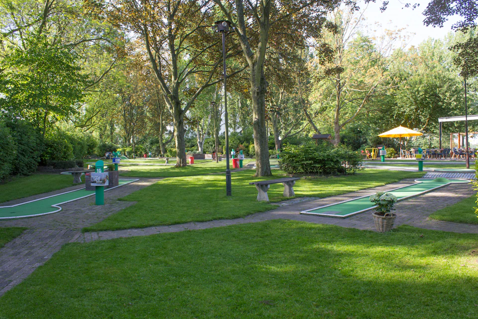 A quiet, sunlit park with mini golf courses, green grass, tall trees, benches, trash bins, and a yellow umbrella in the distance.