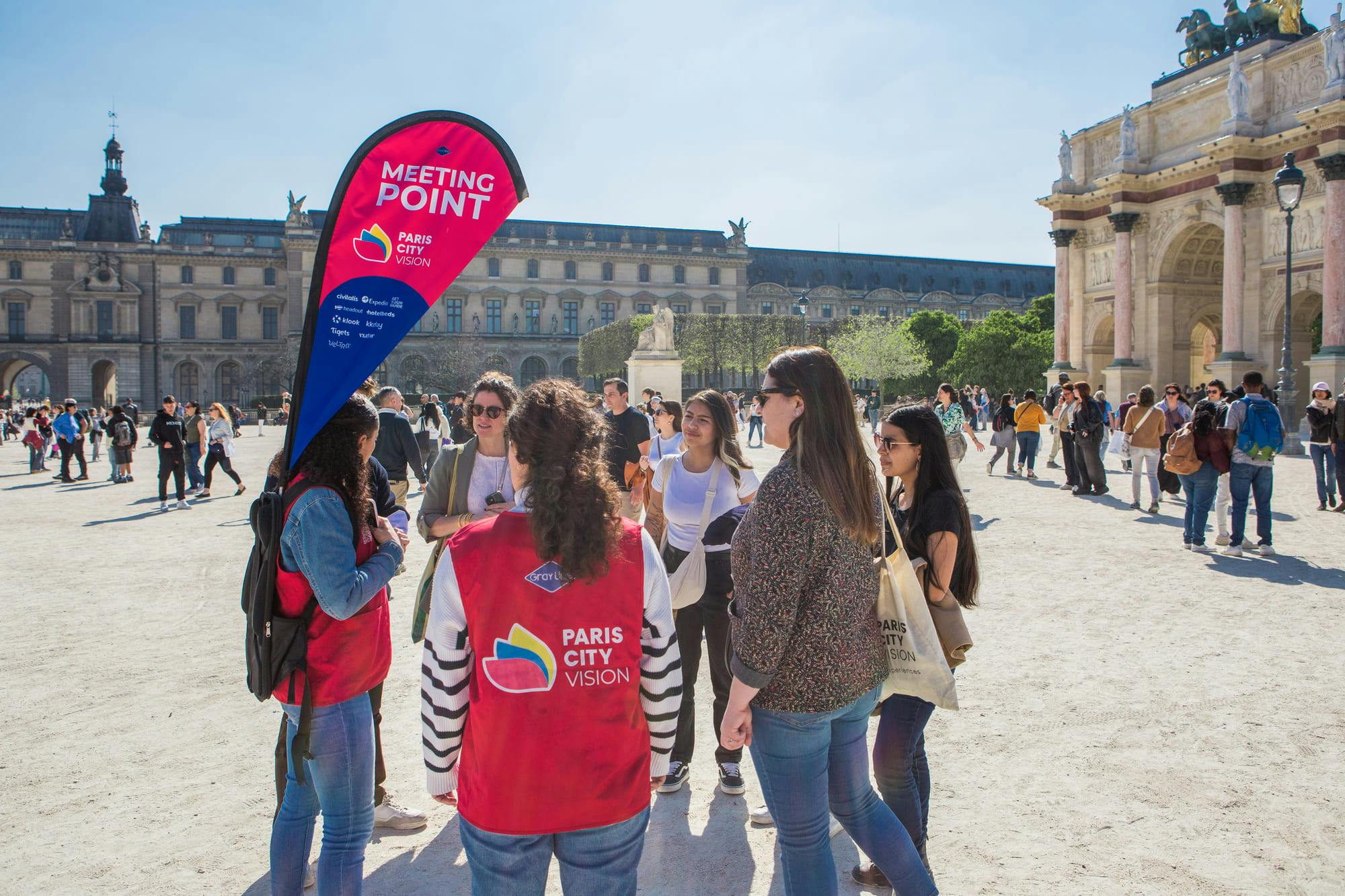 Eine Gruppe von Menschen steht in der Nähe eines "Meeting Point"-Schildes für "Paris City Vision" auf einem sonnigen Platz mit historischen Gebäuden.