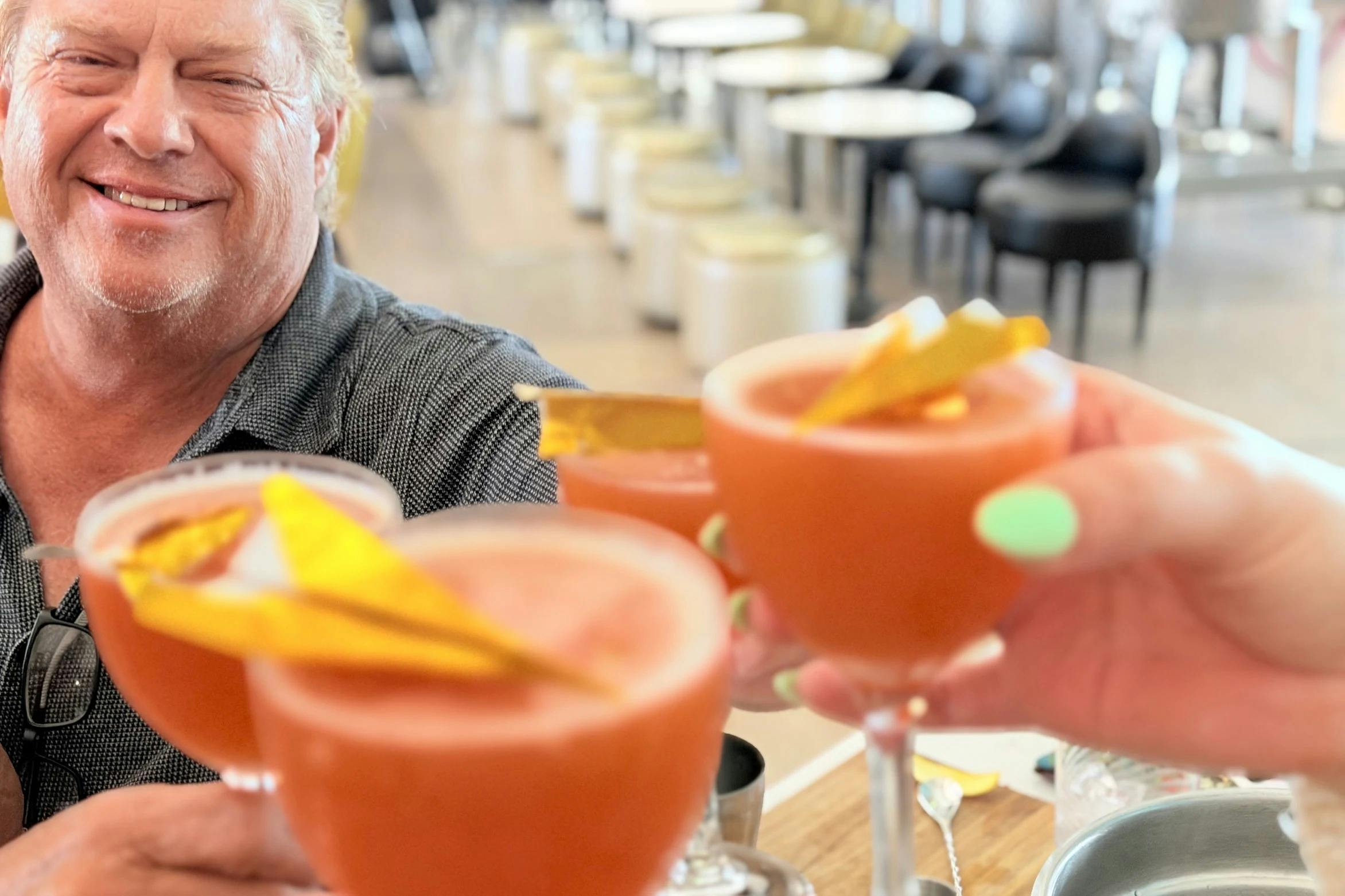 A person smiling while holding a cocktail glass with decorative garnishes, with similar glasses held in the foreground.