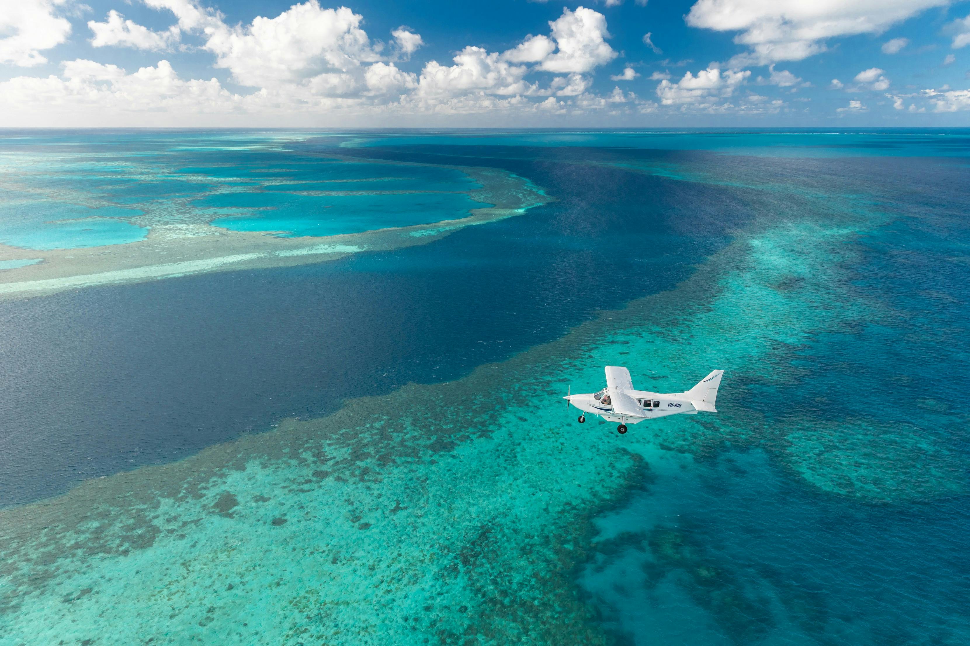 A small airplane flying over a vibrant blue ocean with visible reefs and patches under a partly cloudy sky.
