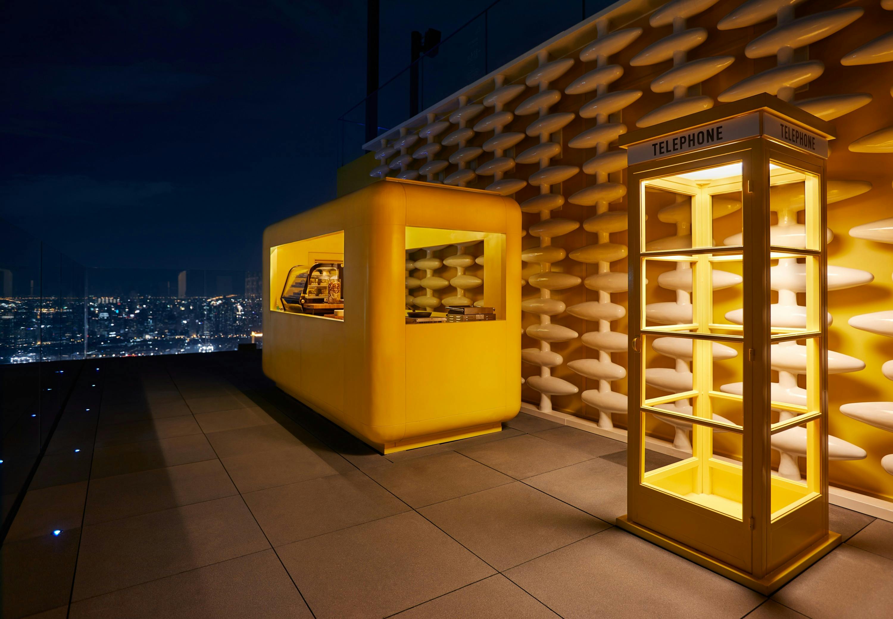 A yellow kiosk and phone booth on a rooftop at night, with a textured wall and city lights in the background.