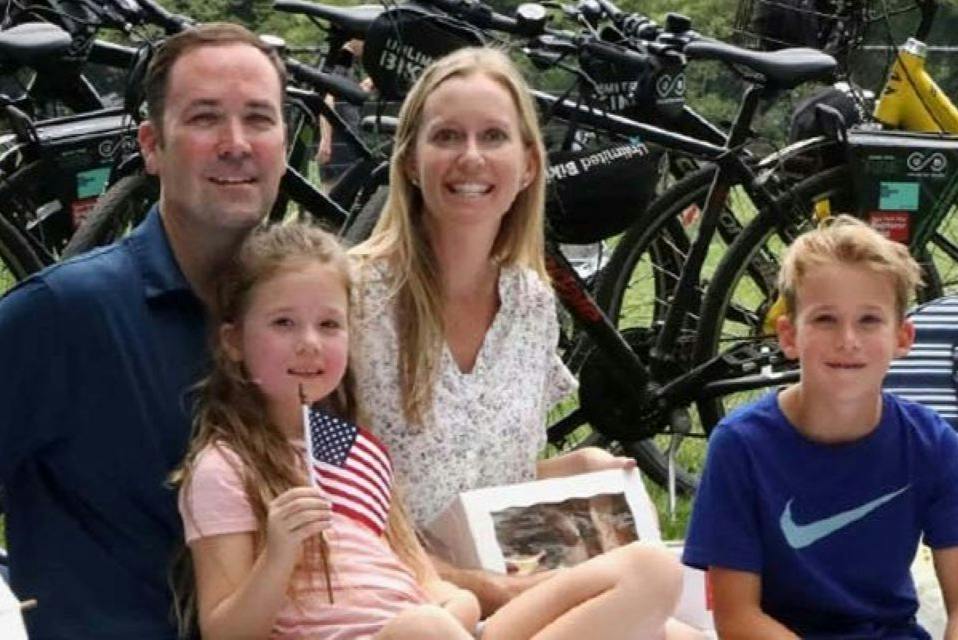 A group of people sitting outdoors near bicycles, with a young girl holding an American flag and others smiling at the camera.
