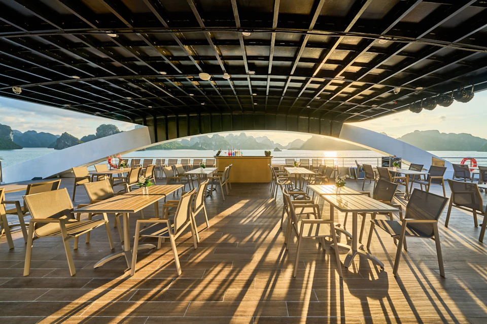 Outdoor dining area on a deck with wooden tables and chairs under a black pergola, overlooking a scenic, sunlit body of water.