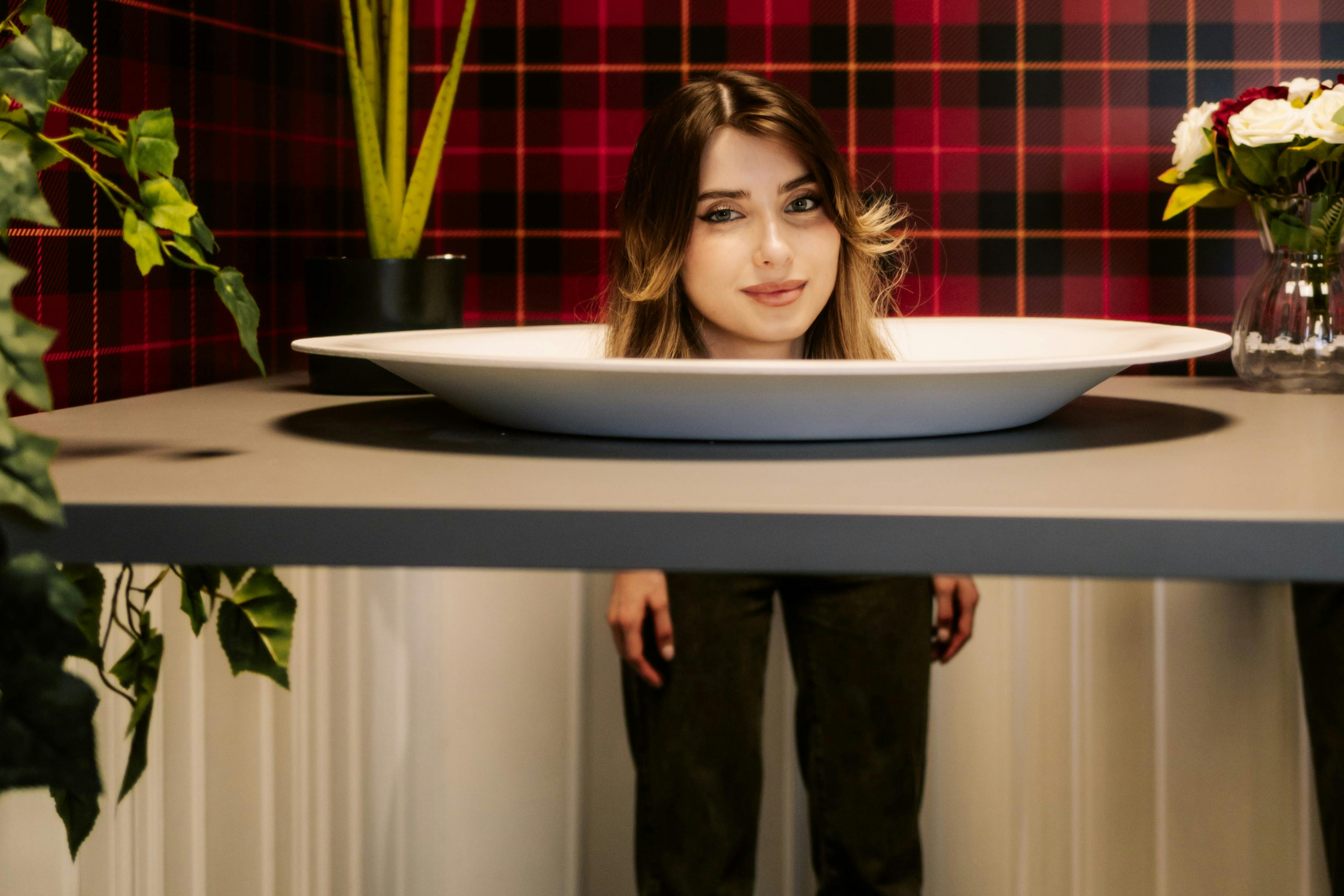 En person verkar ha huvudet på en tallrik som en del av en optisk illusion, omgiven av växter och en röd rutig bakgrund.