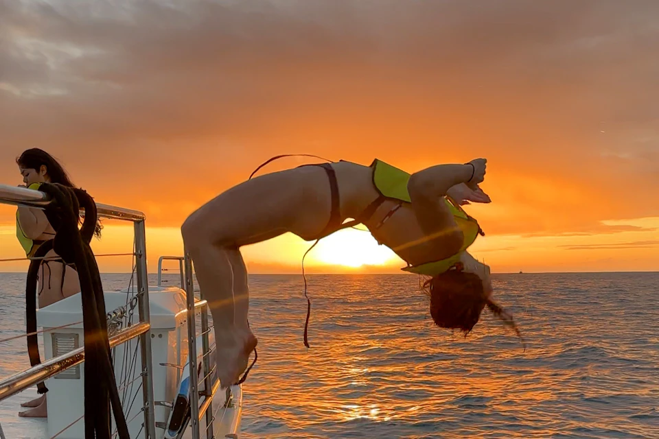 Sunset splash! Flip off a catamaran into glowing orange waters, framed by a fiery Hawaiian sky.