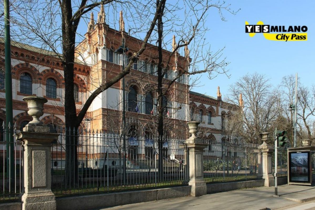 Ein historisches Backsteingebäude mit Rundbogenfenstern, umgeben von einem schwarzen Eisenzaun und kahlen Bäumen.