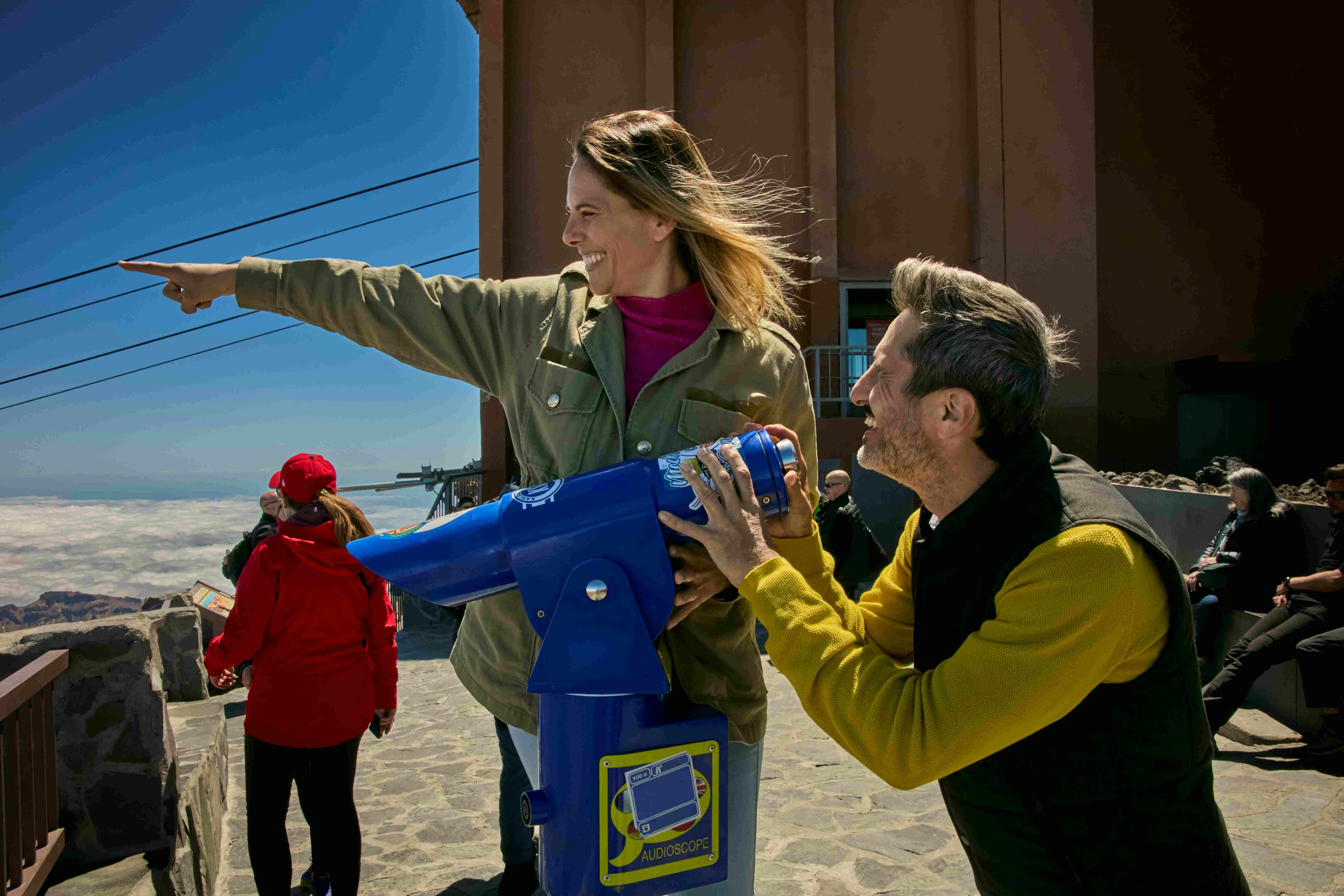 Un home i una dona somrient mentre utilitzen un telescopi blau en un dia assolellat, amb una altra persona al fons.