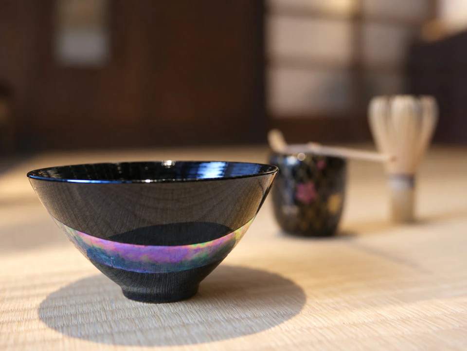 A black bowl with an iridescent stripe on a mat, accompanied by a whisk and a cup in a softly lit setting.
