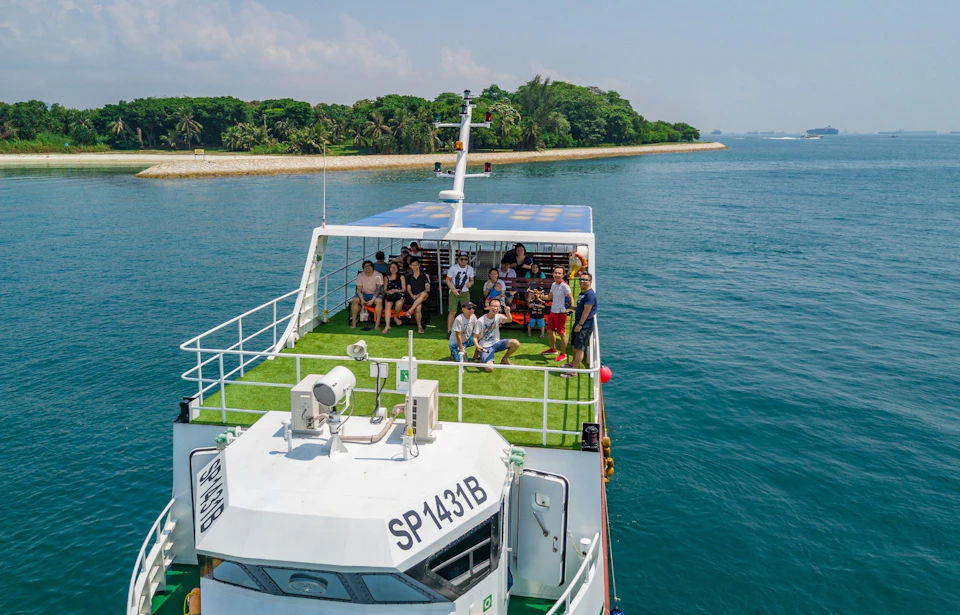 People gathered on the upper deck of a boat with a green floor, near an island with trees, under a clear sky.