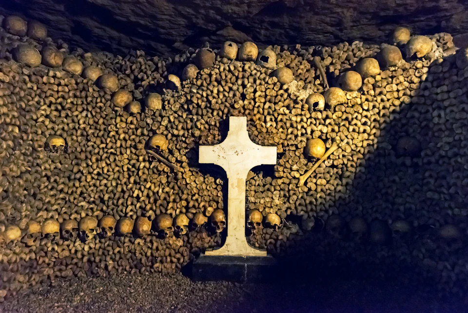 A white cross is centered in front of a wall made of arranged skulls and bones in a dimly lit underground setting.