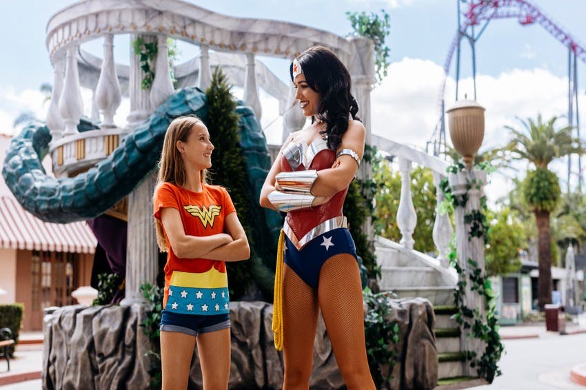 A young girl in a Wonder Woman t-shirt smiles at a woman dressed as Wonder Woman in a theme park with a decorative bridge behind them.